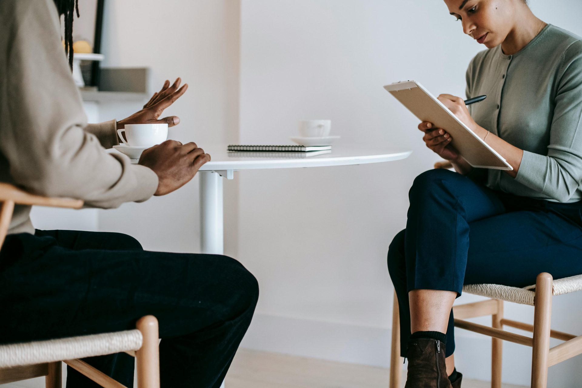 Crop ethnic female psychotherapist in formal clothes writing notes in clipboard while listening to black male patient complains in light studio